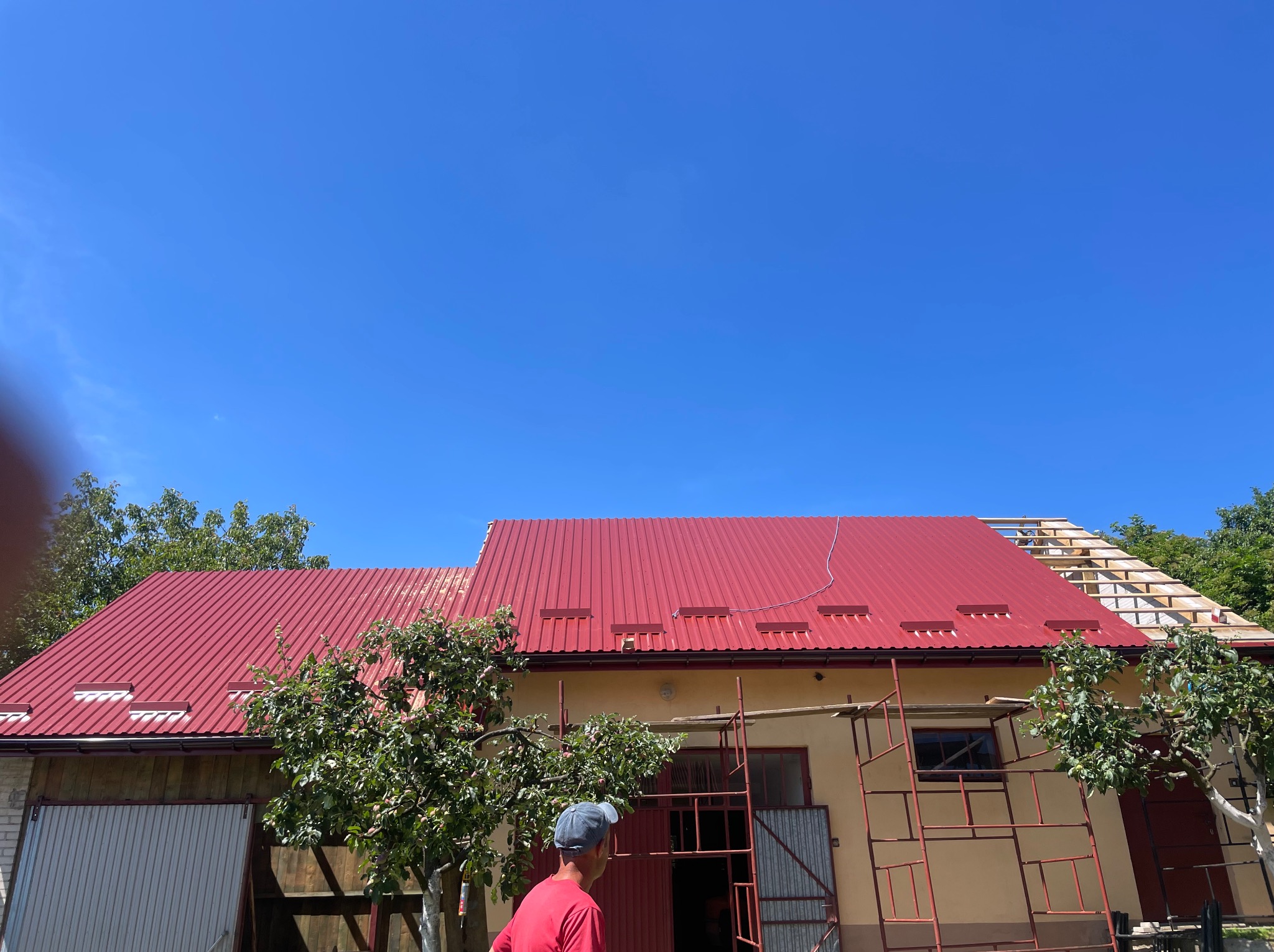 Czerwony blaszany dach na budynku gospodarczym w trakcie prac, widoczne rusztowanie i fragment drewnianej konstrukcji dachu w budowie, na pierwszym planie mężczyzna w czerwonej koszulce i czapce.