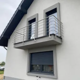 Nowoczesny balkon z balustradą ze stali nierdzewnej, widok z dołu na szarą elewację domu w deszczowy dzień, z widocznym skuterem i klimatyzatorem na dole.