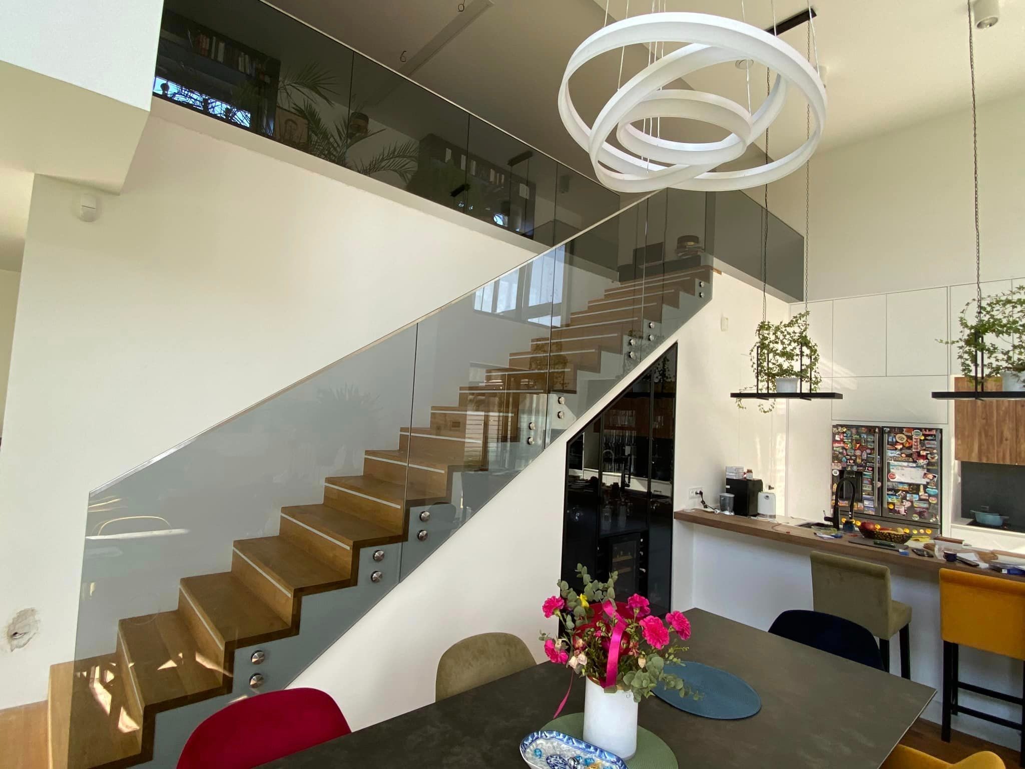 Nowoczesne schody drewniane z balustradą szklaną, widok z dołu, oświetlenie sufitowe w kształcie pierścieni, kwiaty na stole w jadalni