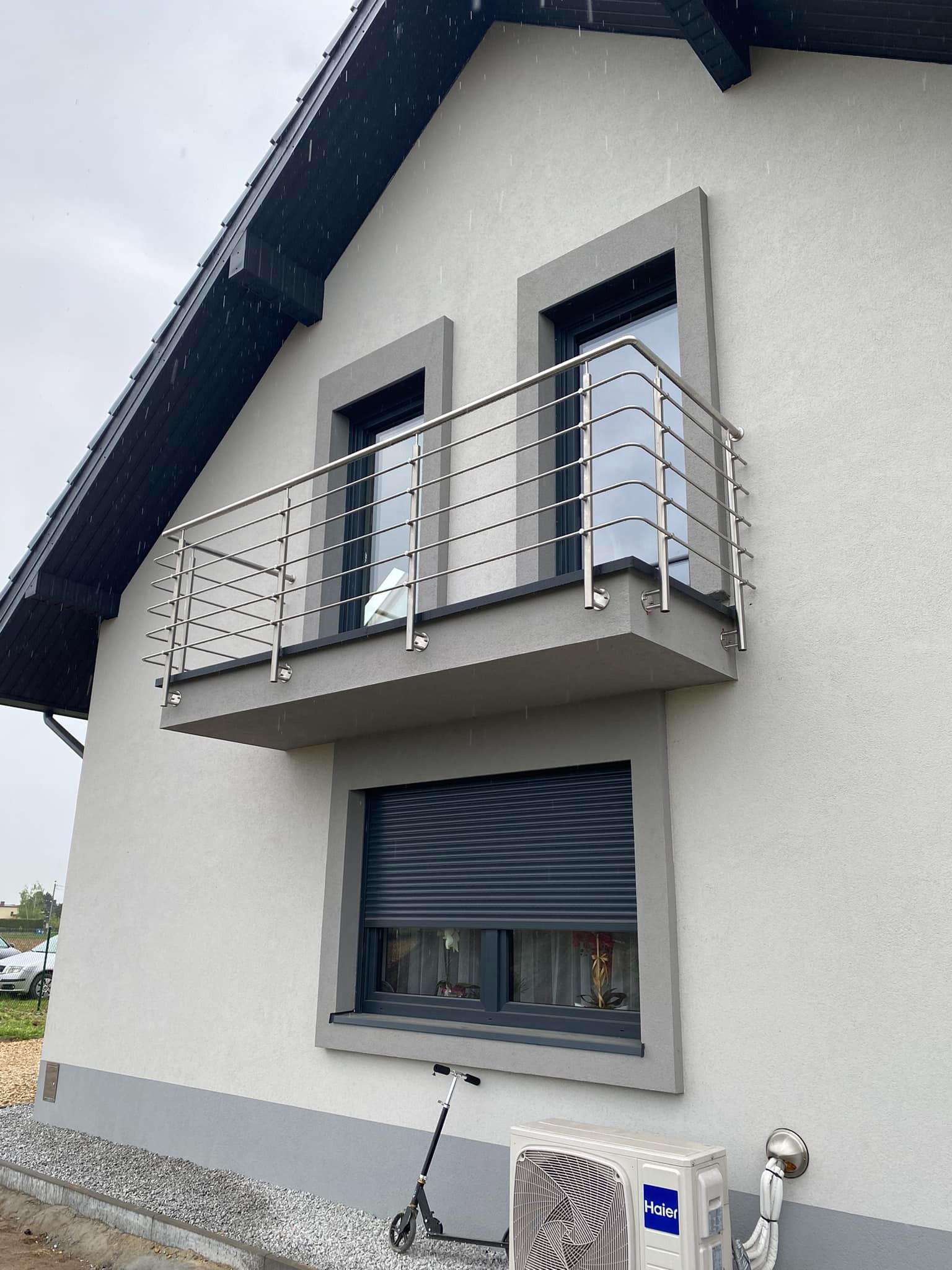Nowoczesny balkon z balustradą ze stali nierdzewnej, widok z dołu na szarą elewację domu w deszczowy dzień, z widocznym skuterem i klimatyzatorem na dole.