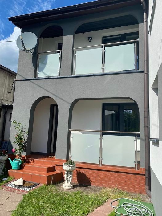 Dwupiętrowy dom z szarą elewacją i balkonami z balustradami ze szkła matowego i stali nierdzewnej, widoczna antena satelitarna na elewacji, czerwone schody wejściowe i fragment ogrodu.