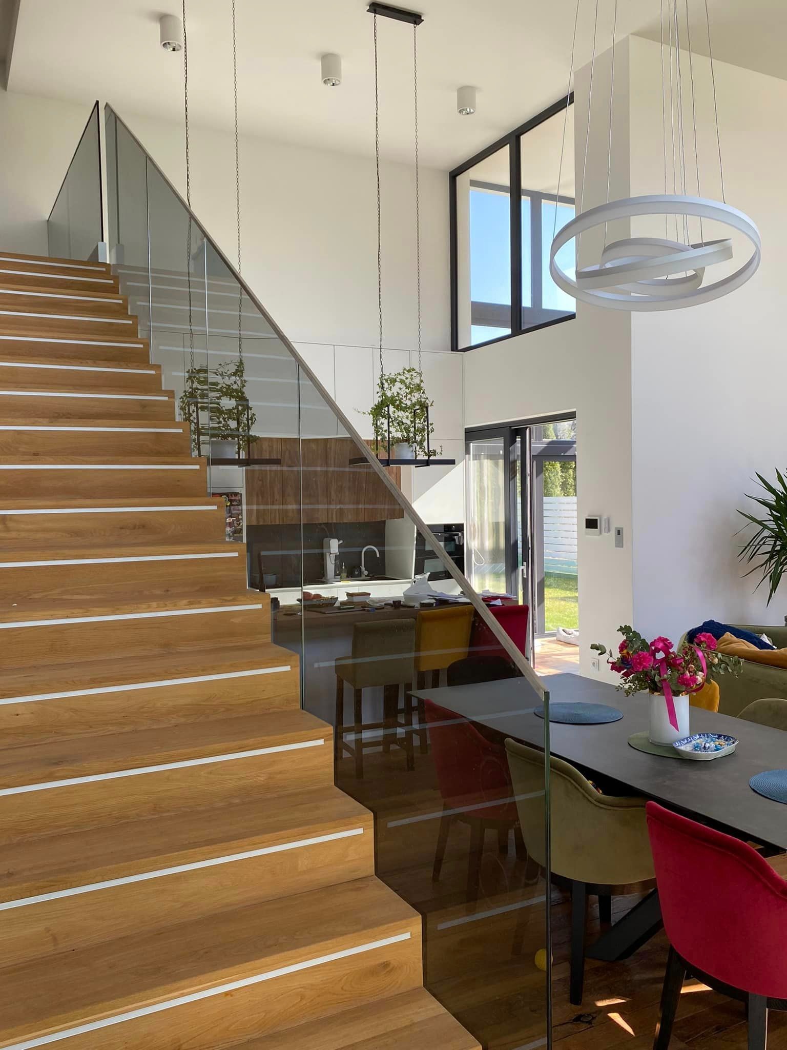Wnętrze domu z drewnianymi schodami z białymi paskami antypoślizgowymi, zabezpieczonymi szklaną balustradą, widok na jadalnię i kuchnię z wiszącymi roślinami.