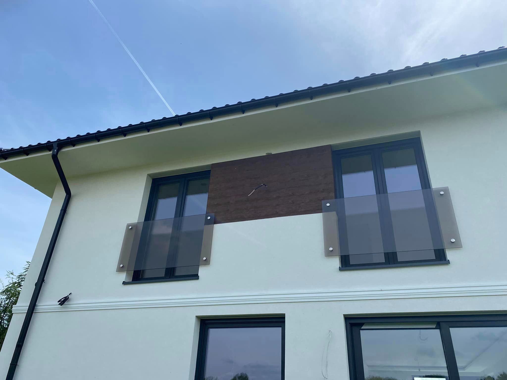 Elewacja budynku z dwoma balkonami zabezpieczonymi szkłem mocowanym na srebrnych śrubach, widok z dołu na tle błękitnego nieba z śladami po samolocie.