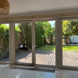 Szerokie, przesuwne okna tarasowe z widokiem na drewniany taras i zadbany ogród, jasne wnętrze z beżowymi ścianami, widoczna lampa z wikliny na suficie.