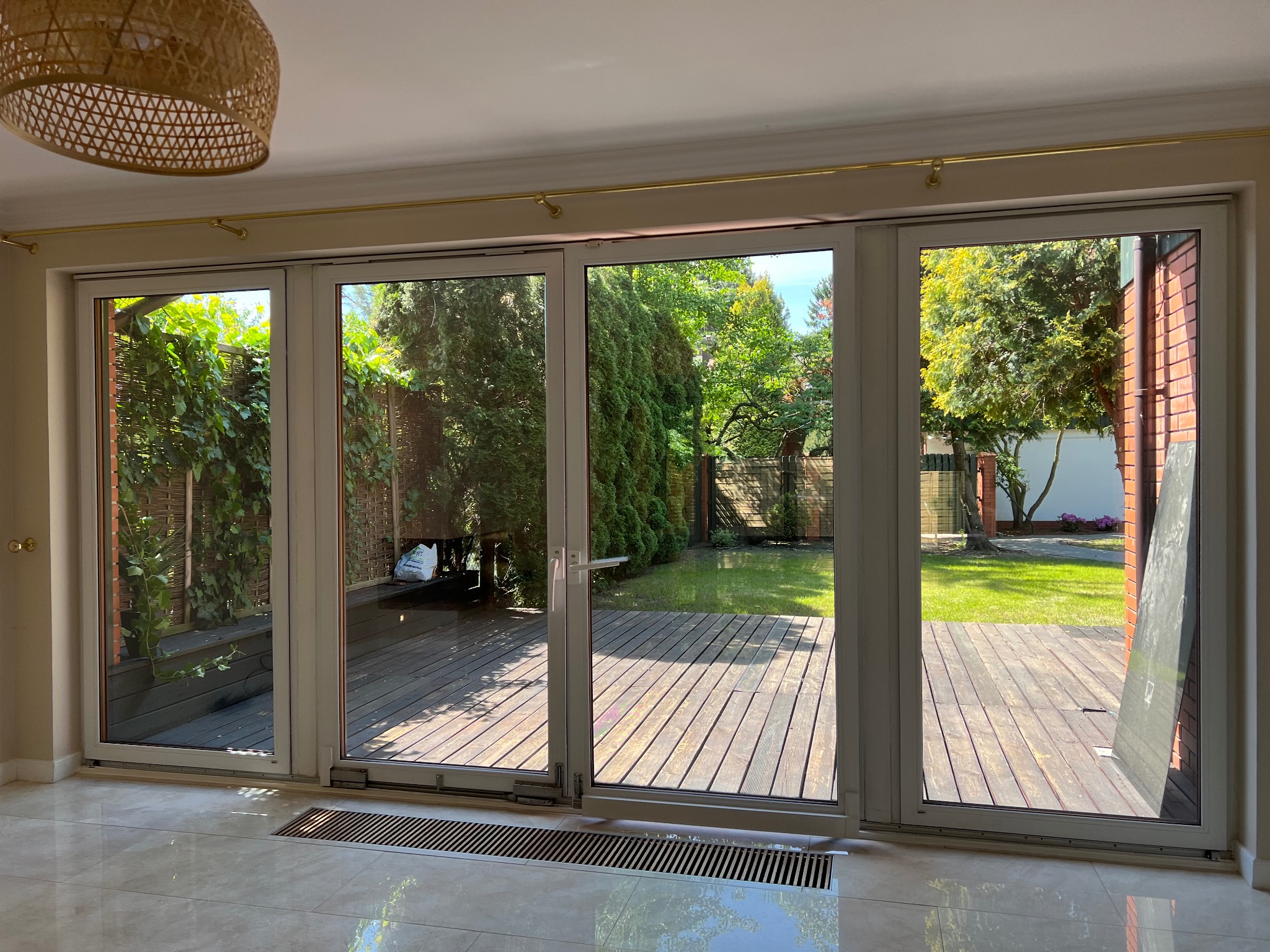 Szerokie, przesuwne okna tarasowe z widokiem na drewniany taras i zadbany ogród, jasne wnętrze z beżowymi ścianami, widoczna lampa z wikliny na suficie.