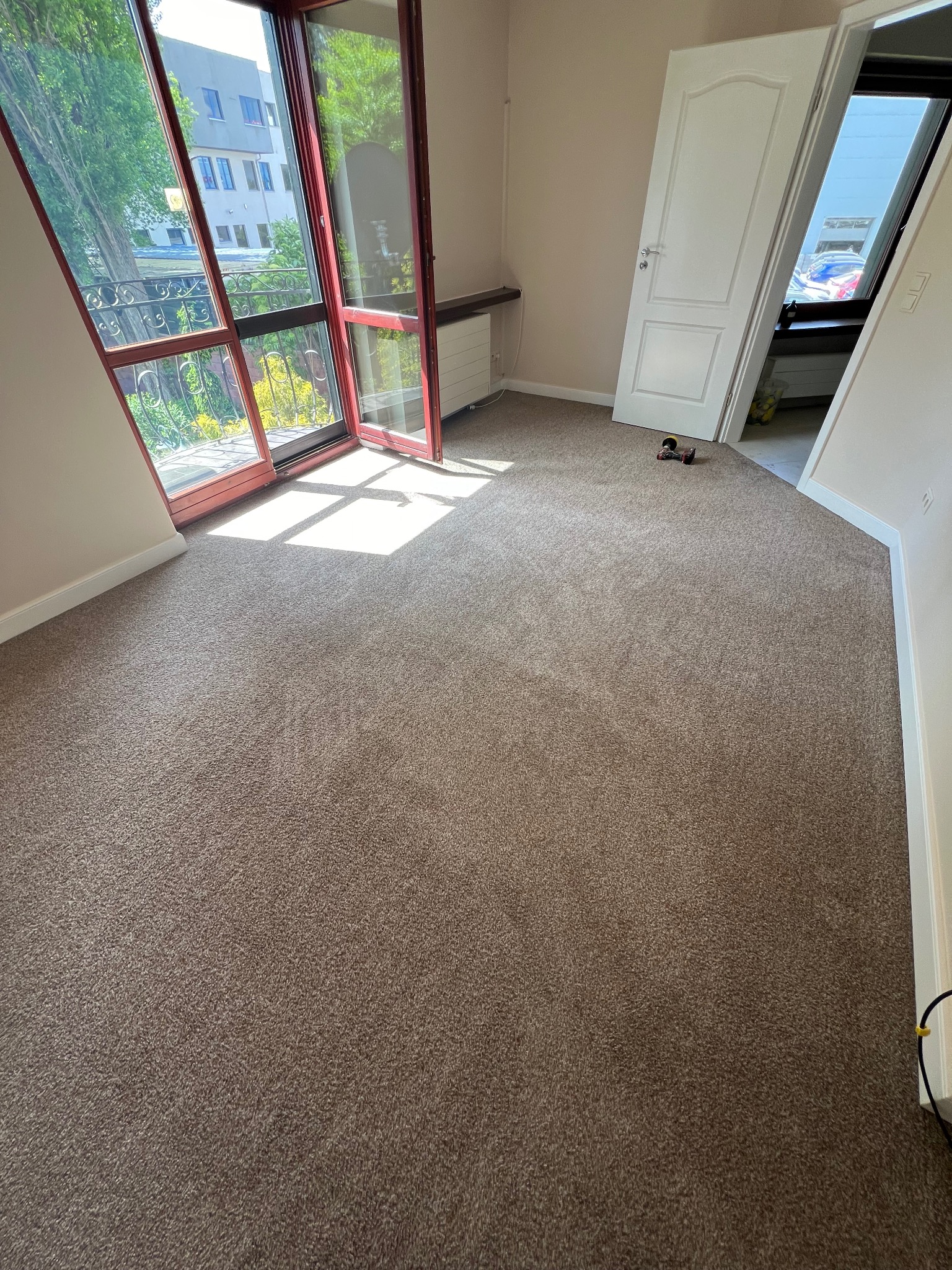 Brązowy dywan w pustym pokoju z otwartymi drzwiami balkonowymi i białymi drzwiami wewnętrznymi, widok na zieleń za oknem.