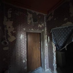 FM Fix Me - Pokój w trakcie przygotowania do remontu: zerwane tapety, widoczne ślady po kleju, drewniane drzwi, fragment sufitu w kolorze pomarańczowym, odsłonięta instalacja elektryczna i fragment zasłony...