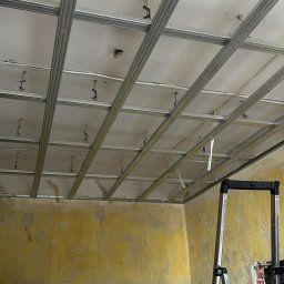 FM Fix Me - Metalowa konstrukcja sufitu podwieszanego, widok z dołu, żółte ściany w tle, drabina z narzędziami po prawej stronie.