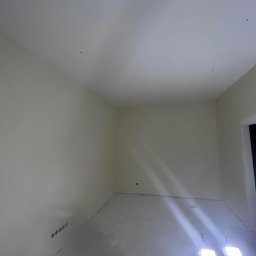 FM Fix Me - Wnętrze pokoju w trakcie remontu, ściany i sufit świeżo pomalowane na jasny kolor, widoczne punkty po szpachlowaniu, na podłodze rozłożony wałek malarski, oświetlone dwoma halogenami.