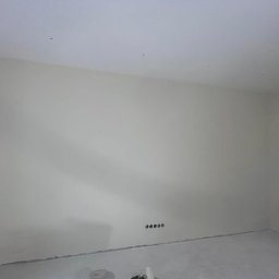 FM Fix Me - Wnętrze pokoju po świeżym szpachlowaniu ścian i sufitu, widoczne gniazdka elektryczne w dolnej części ściany, podłoga zabezpieczona folią malarską.