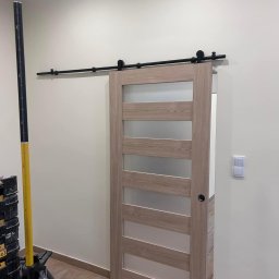 FM Fix Me - Zamontowane przesuwne drzwi drewniane z matowym szkłem, zawieszone na czarnym systemie szynowym, obok stoi żółty laserowy niwelator na trójnogu, widoczny fragment podłogi i ściany z włącznikiem.