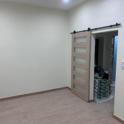 FM Fix Me - Wnętrze pokoju z drewnianą podłogą i przesuwnymi drzwiami z mlecznego szkła; widoczne worki z zaprawą budowlaną w przejściu do kolejnego pomieszczenia.