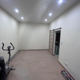 FM Fix Me - Pusty pokój po remoncie z nową podłogą, widoczne worki z materiałami budowlanymi w głębi oraz maszyna do ćwiczeń w rogu.