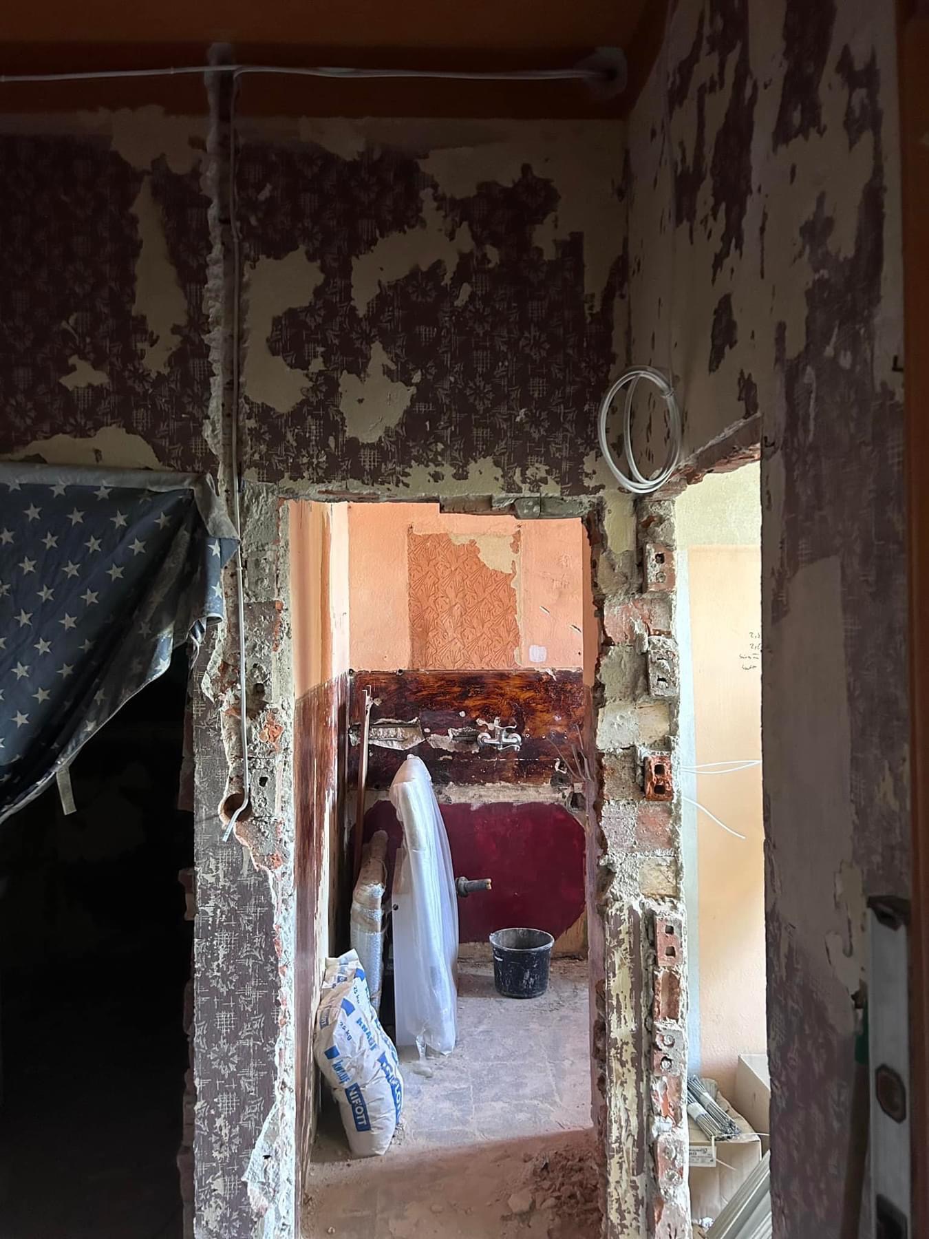 Demontaż starej okładziny ściennej w trakcie remontu z widocznymi cegłami i pozostałościami tapety, worki z zaprawą budowlaną na pierwszym planie, widoczne przewody elektryczne.