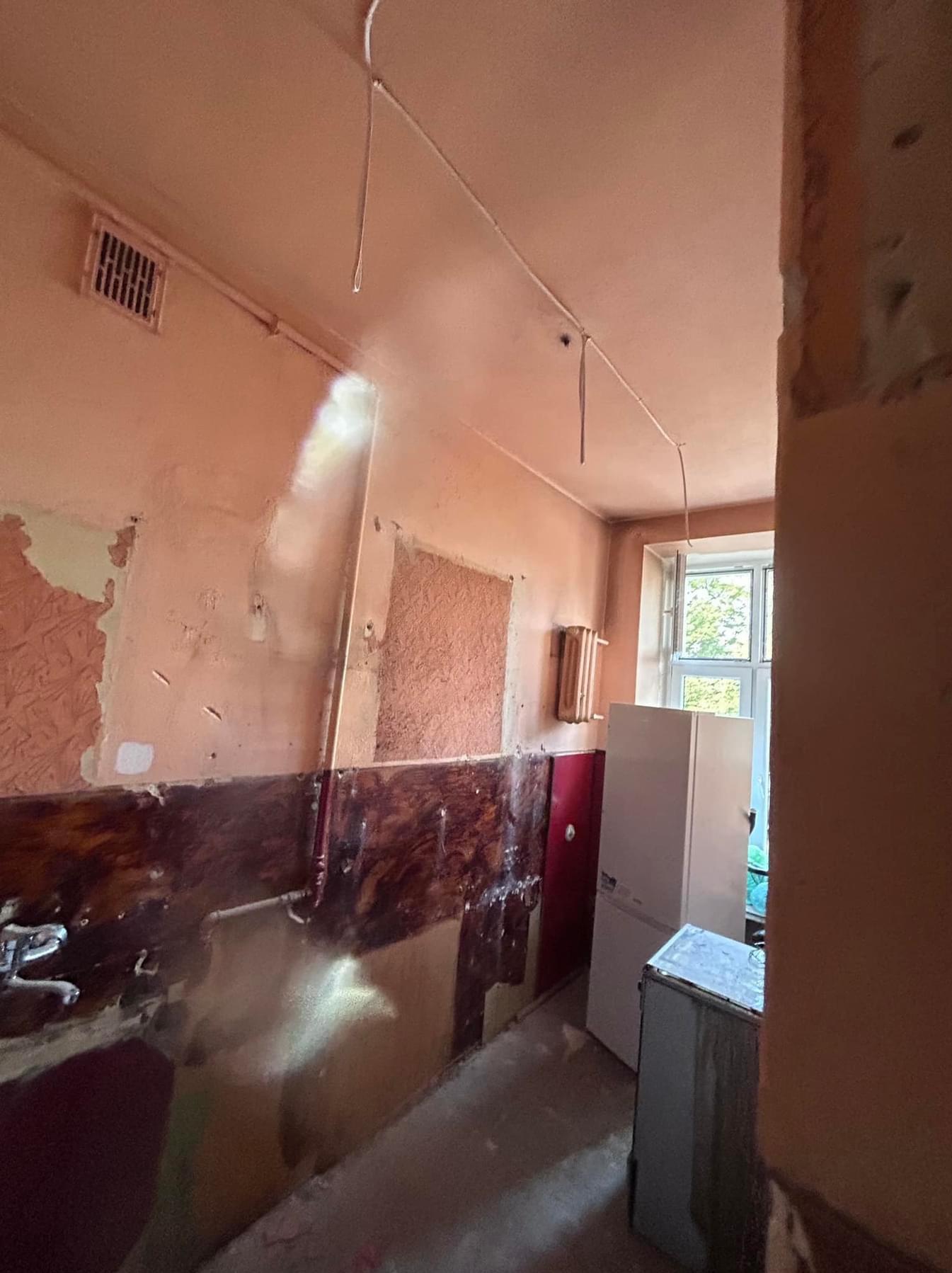 Kuchnia w trakcie remontu: ściany z częściowo usuniętą farbą, odsłonięte instalacje elektryczne, widoczny fragment lodówki i okna.