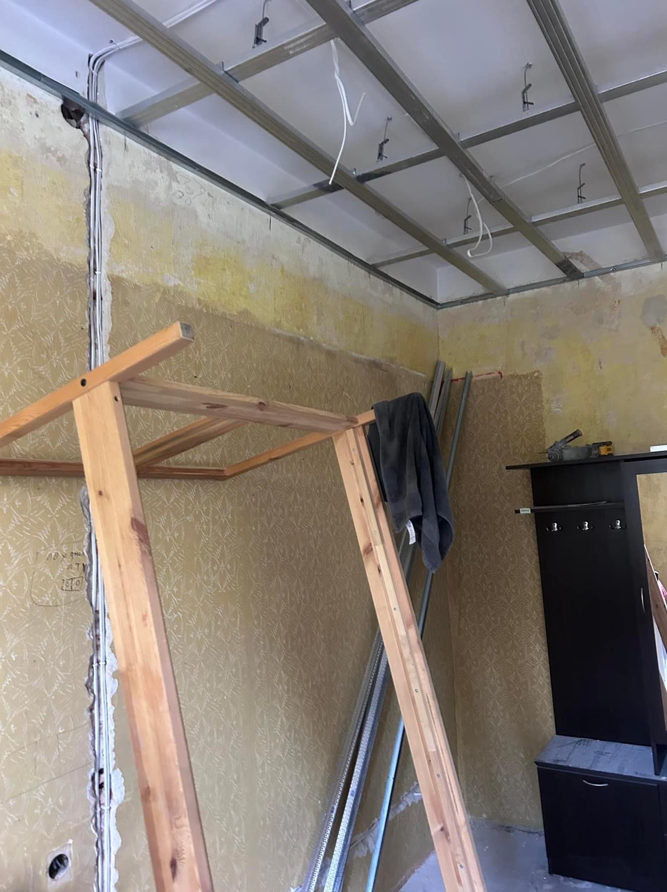 Wnętrze w trakcie remontu z widoczną konstrukcją metalową pod sufit podwieszany, odsłoniętą instalacją elektryczną w ścianie, starą tapetą, drewnianym stelażem i elementami garderoby w tle.
