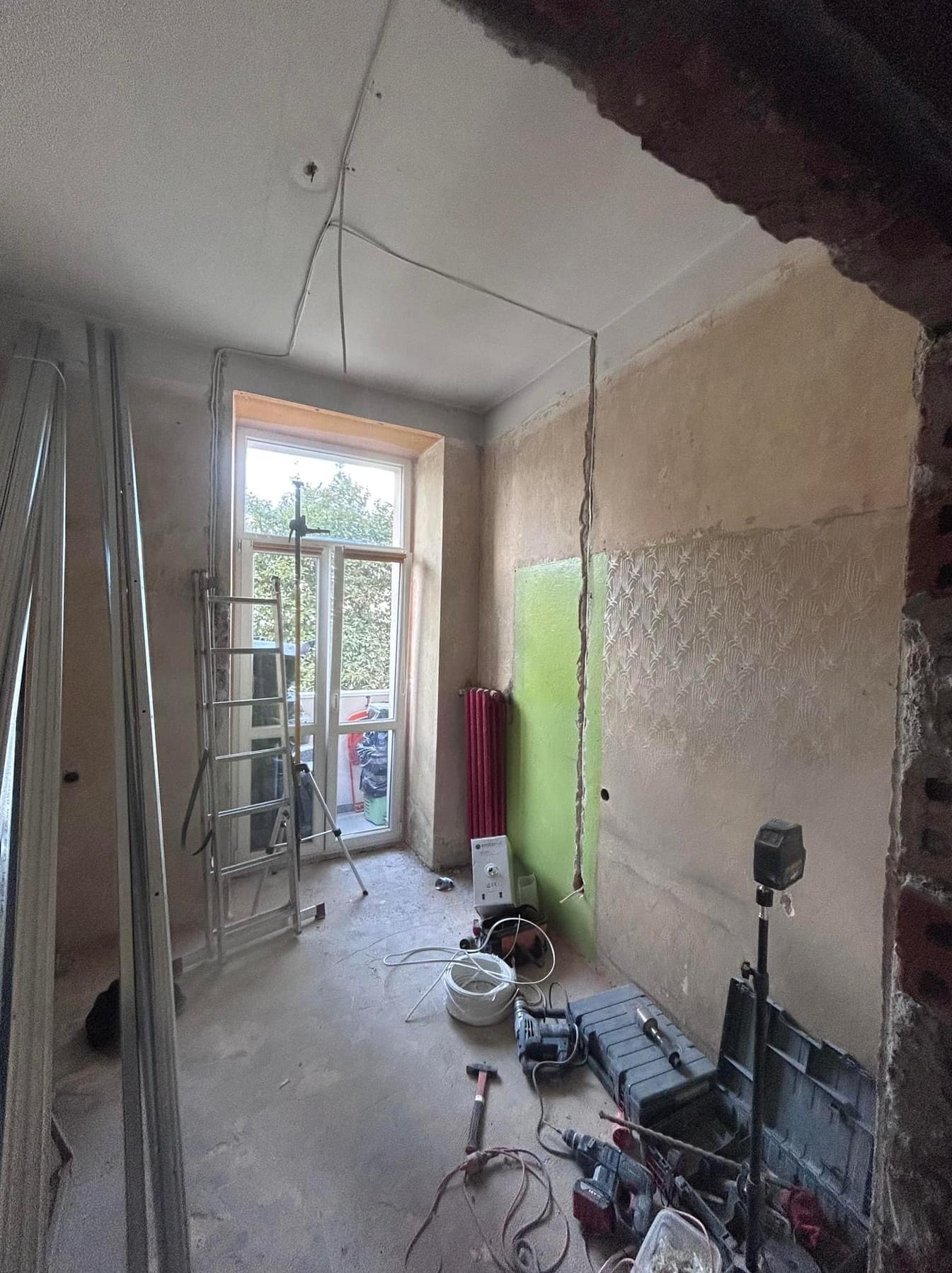 Pokój w trakcie remontu: widoczne profile do płyt karton gips, drabina, narzędzia, fragment ściany z pozostałościami tapety i nową instalacją elektryczną.