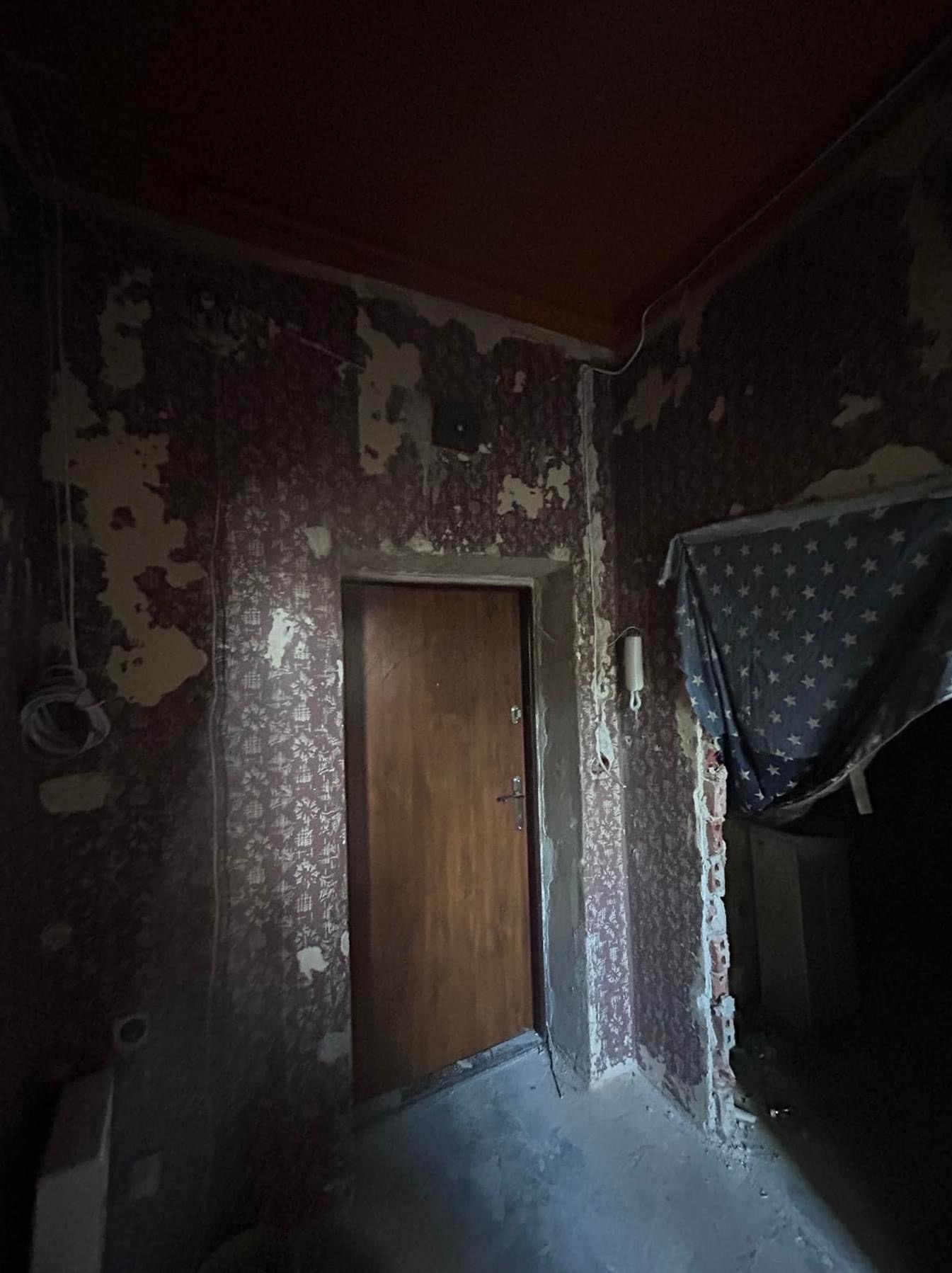 Pokój w trakcie przygotowania do remontu: zerwane tapety, widoczne ślady po kleju, drewniane drzwi, fragment sufitu w kolorze pomarańczowym, odsłonięta instalacja elektryczna i fragment zasłony...