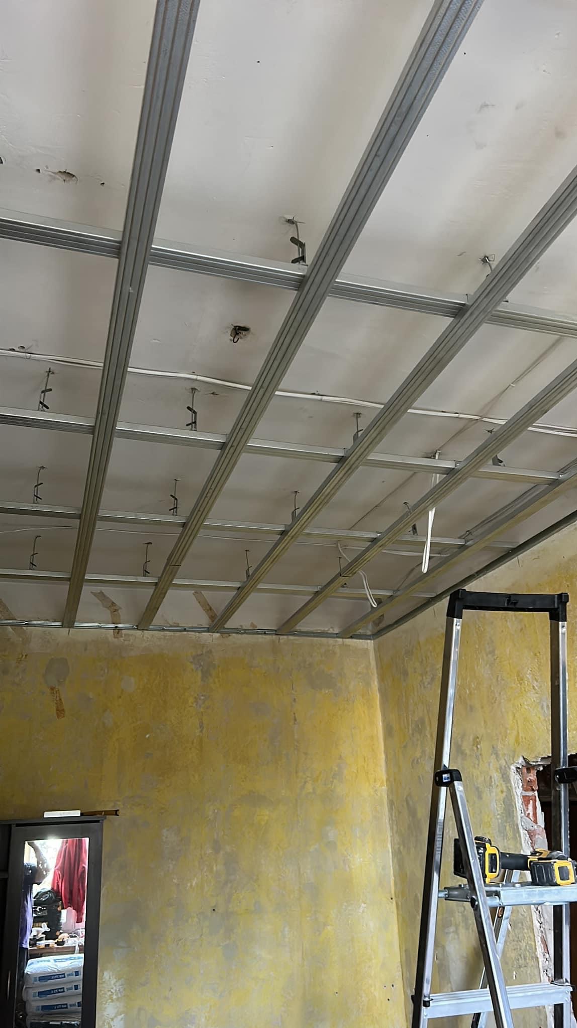 Metalowa konstrukcja sufitu podwieszanego, widok z dołu, żółte ściany w tle, drabina z narzędziami po prawej stronie.