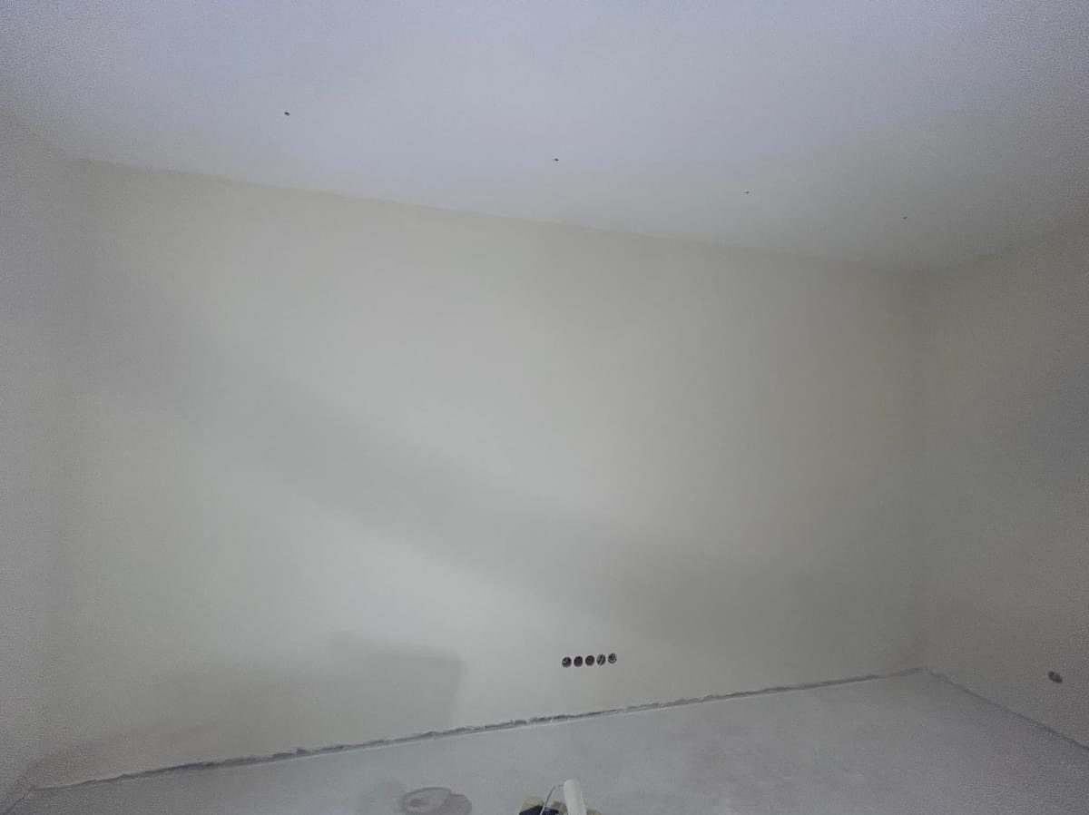 Wnętrze pokoju po świeżym szpachlowaniu ścian i sufitu, widoczne gniazdka elektryczne w dolnej części ściany, podłoga zabezpieczona folią malarską.