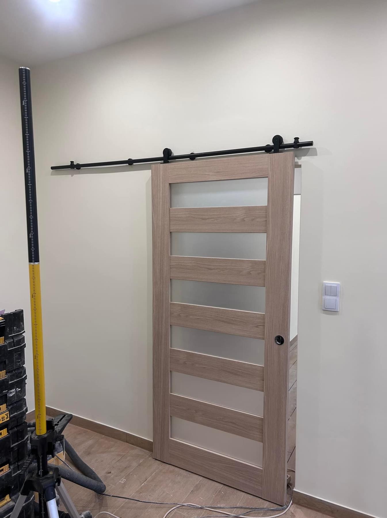 Zamontowane przesuwne drzwi drewniane z matowym szkłem, zawieszone na czarnym systemie szynowym, obok stoi żółty laserowy niwelator na trójnogu, widoczny fragment podłogi i ściany z włącznikiem.