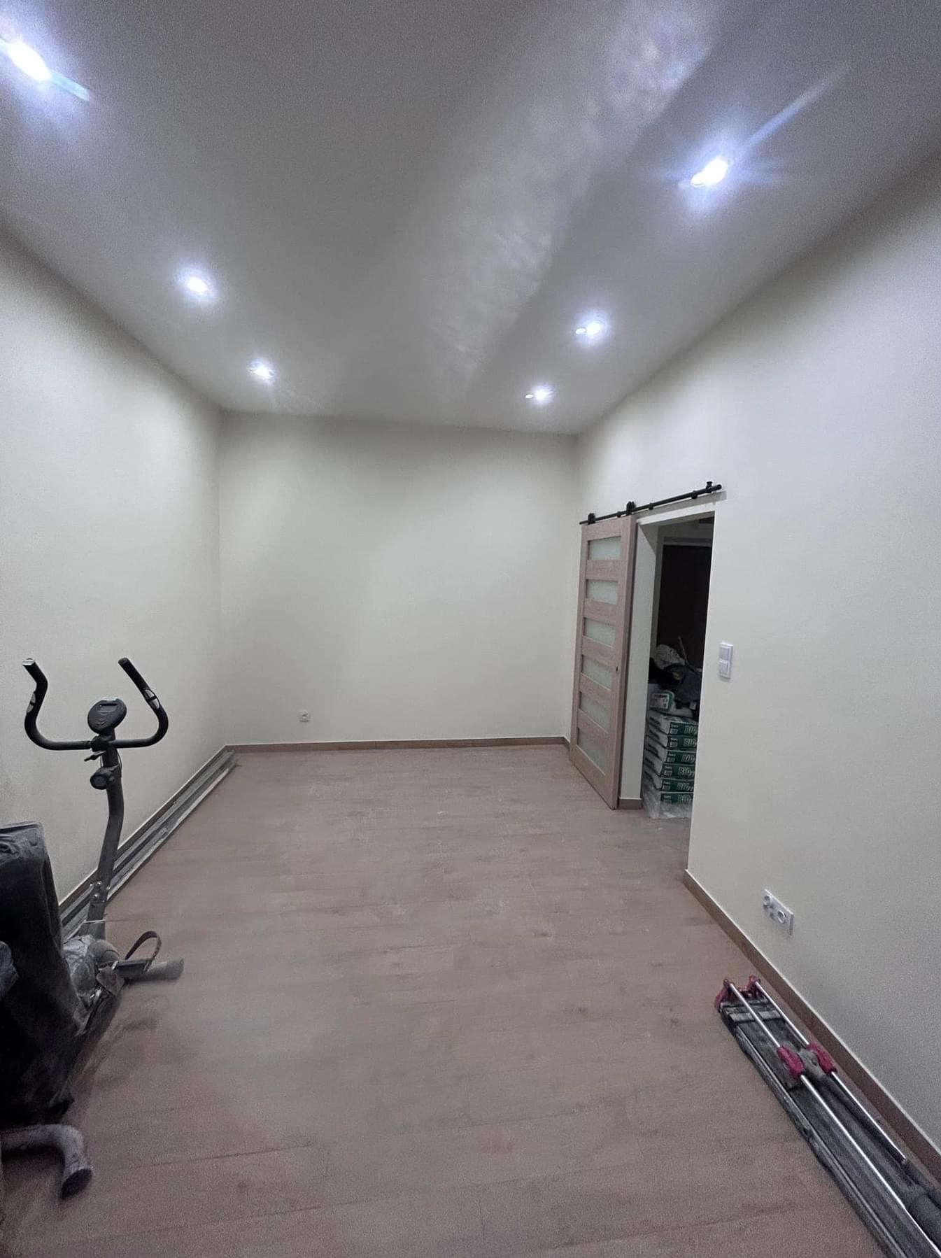 Pusty pokój po remoncie z nową podłogą, widoczne worki z materiałami budowlanymi w głębi oraz maszyna do ćwiczeń w rogu.