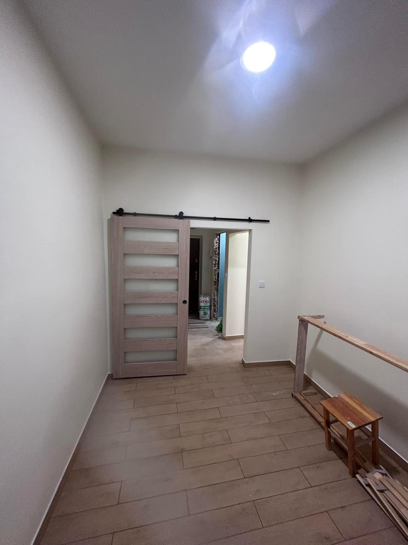 Wnętrze po remoncie: przesuwne drzwi z mlecznym szkłem na czarnej szynie, ułożona podłoga z płytek imitujących drewno, widoczna torba z zaprawą budowlaną w przejściu do innego pomieszczenia.