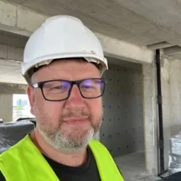 Pracownik w kasku i kamizelce odblaskowej na tle surowego wnętrza budynku w trakcie budowy, widoczne ocieplenie ścian i instalacje.