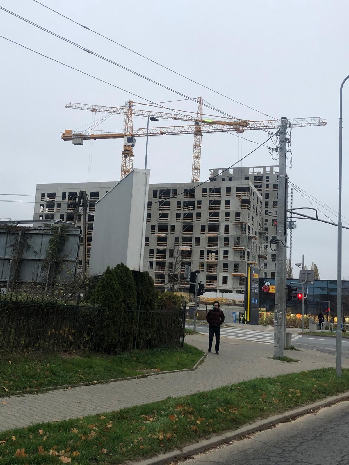 Budynek w stanie surowym otwartym z widocznymi otworami okiennymi, dźwigi budowlane w tle, osoba na chodniku w pobliżu przejścia dla pieszych.