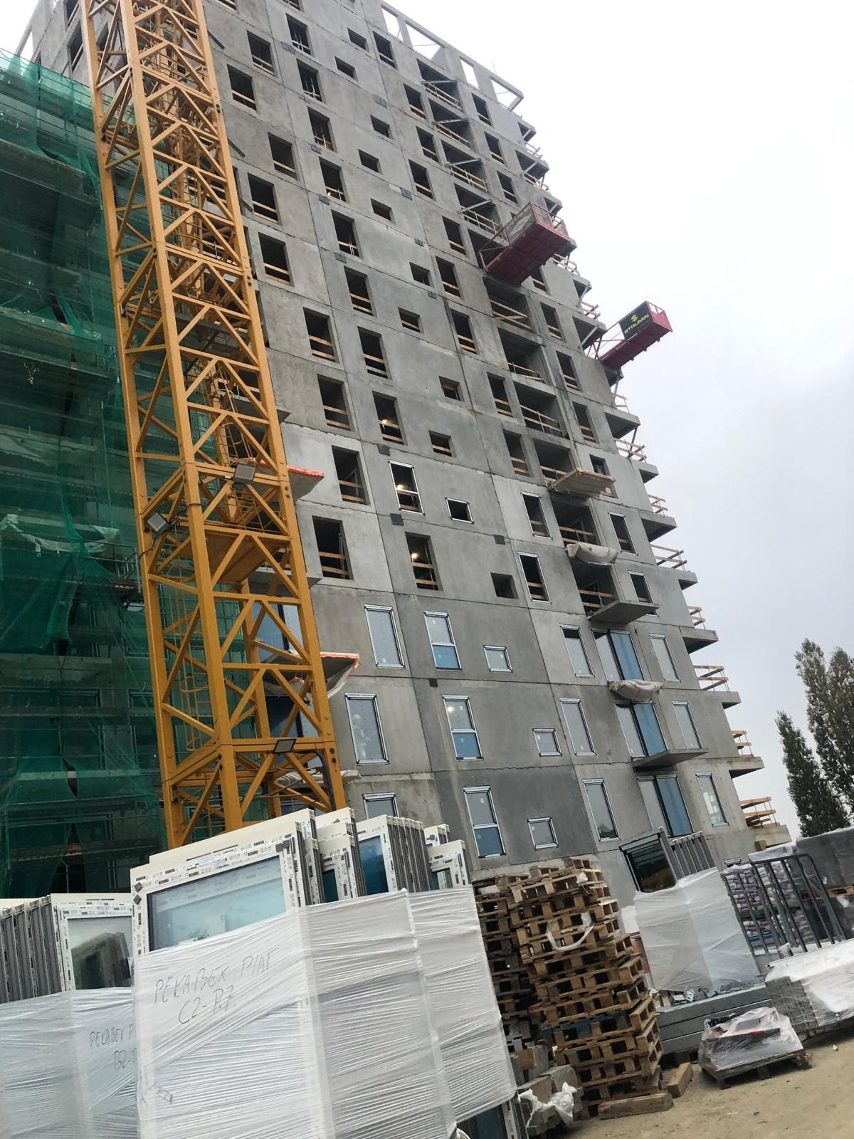 Budynek wielorodzinny w trakcie montażu okien, widoczne dźwigi, rusztowania i okna przygotowane do instalacji, zapakowane w folię ochronną.