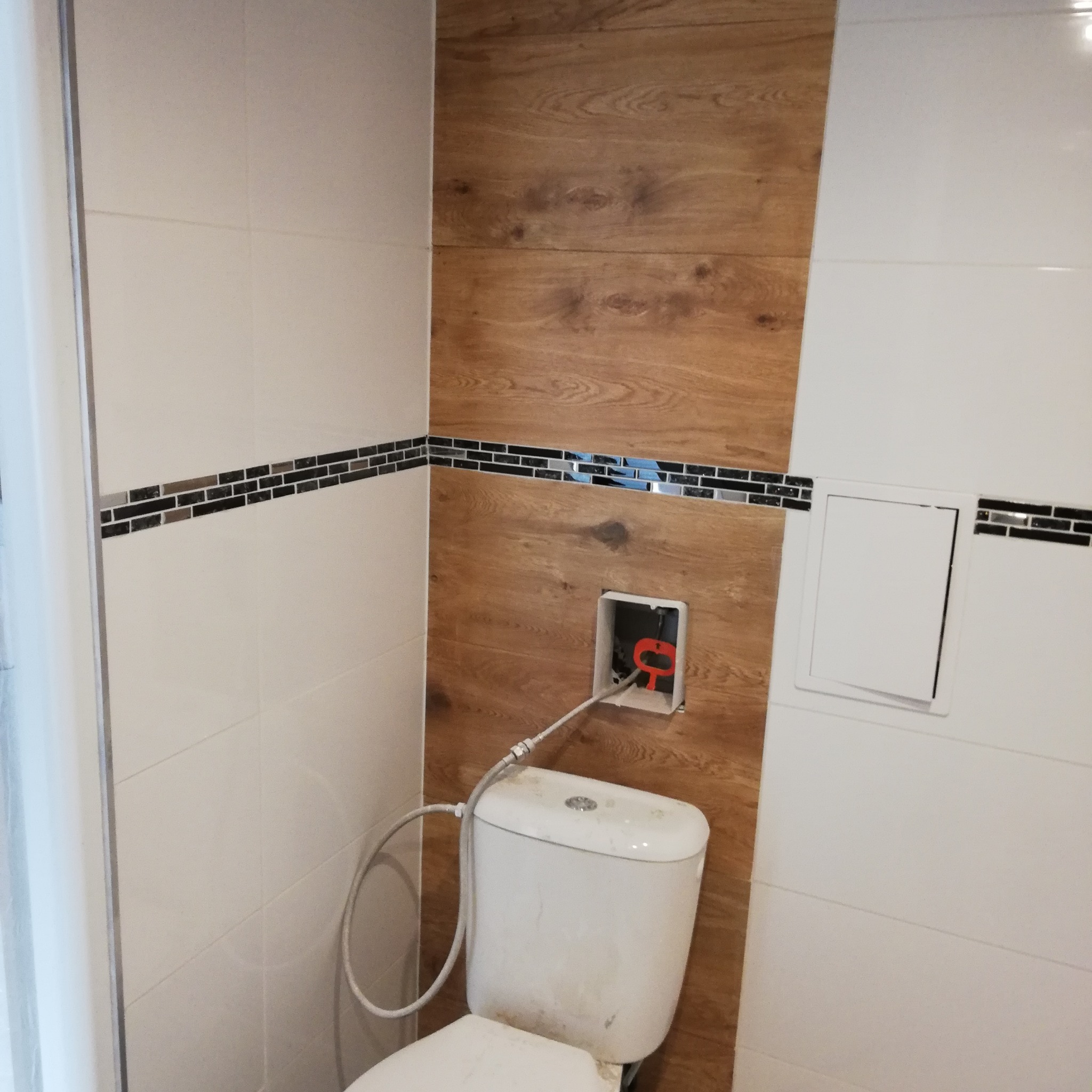 Biała toaleta z podłączeniem do instalacji w trakcie montażu na tle ściany wyłożonej białymi i drewnopodobnymi płytkami z dekoracyjnym paskiem mozaiki.