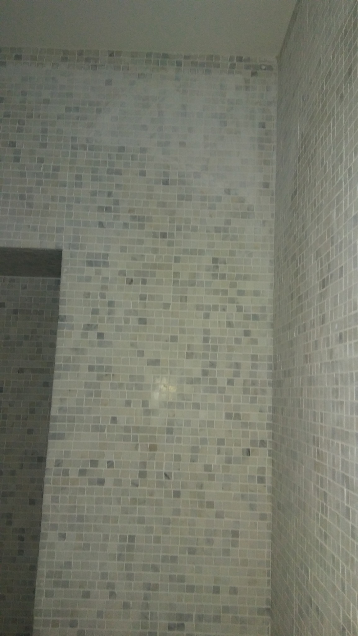 Ściana łazienki wyłożona mozaiką z jasnych, szarych płytek w kształcie małych kwadratów, widoczny narożnik i fragment wnęki.