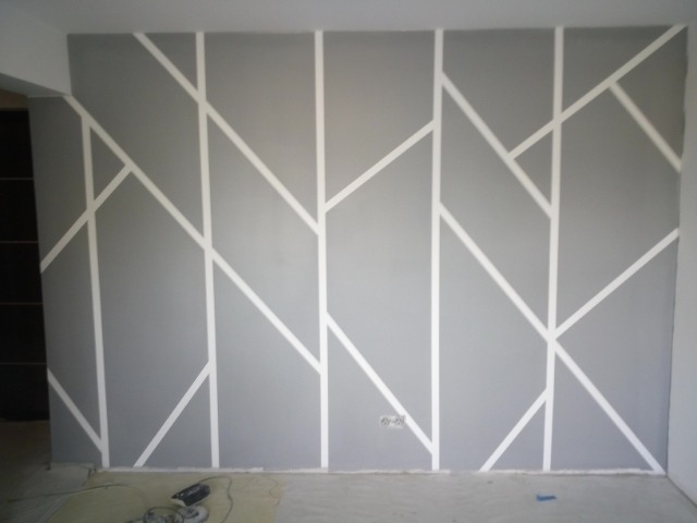 Szara ściana z geometrycznym wzorem z białych pasów w trakcie malowania w Łodzi, widoczne narzędzia malarskie na podłodze.