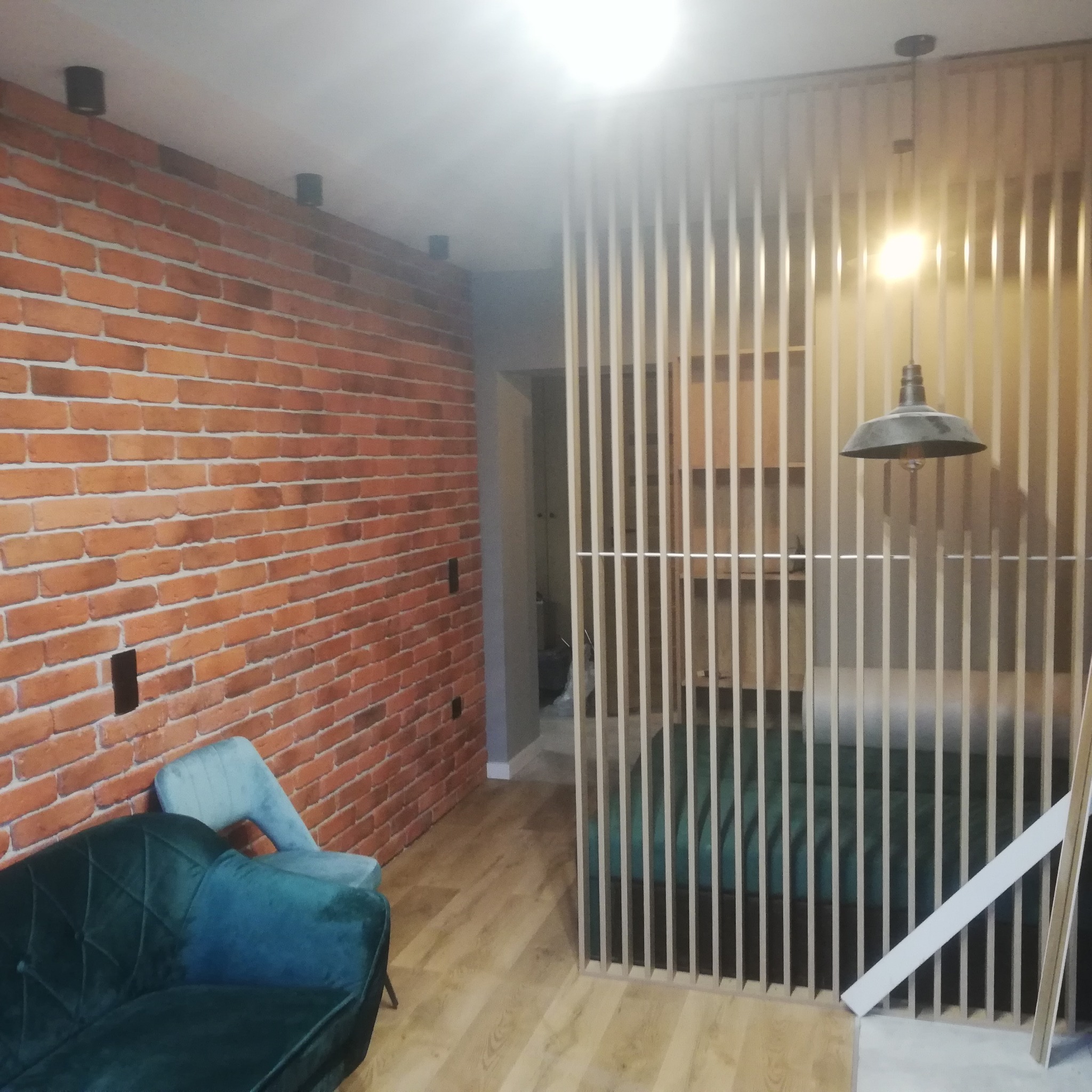 Salon z ceglaną ścianą, drewnianą ażurową ścianką działową, zieloną sofą i lampą wiszącą.