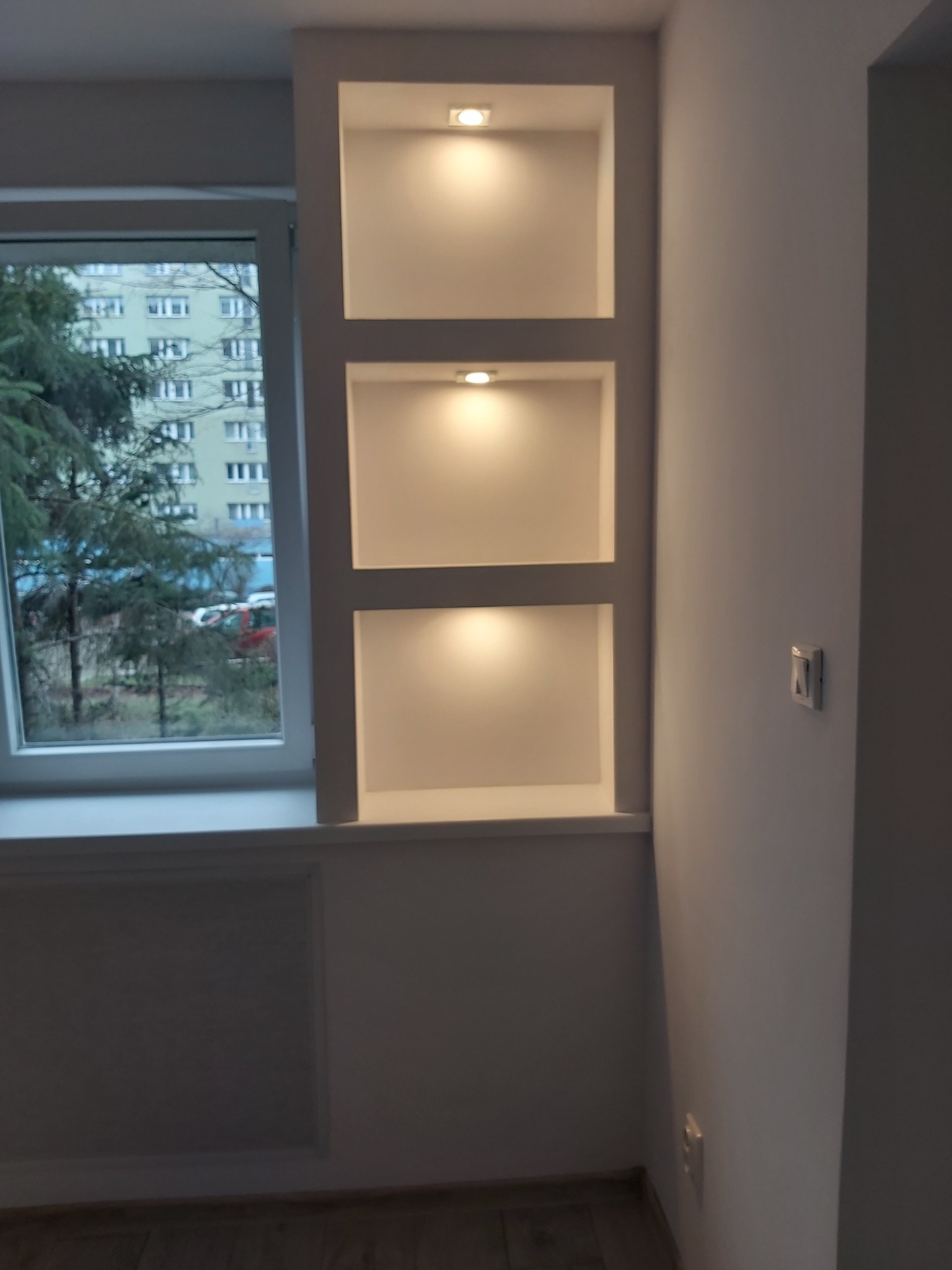 Wnęka ścienna z trzema podświetlanymi półkami obok okna z widokiem na budynek mieszkalny i drzewa.