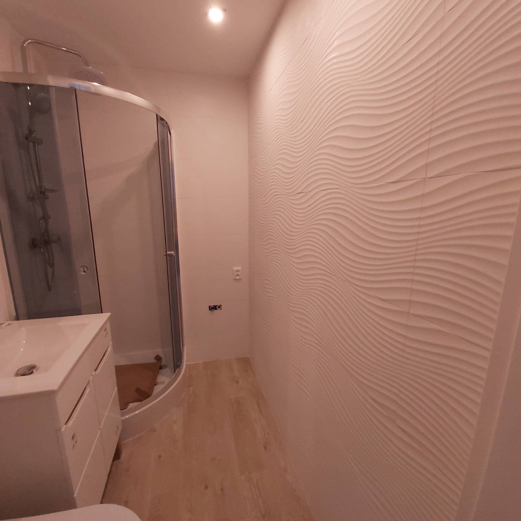 Nowoczesna łazienka z kabiną prysznicową, umywalką z szafką i ścianą wyłożoną białymi płytkami z falistym wzorem.