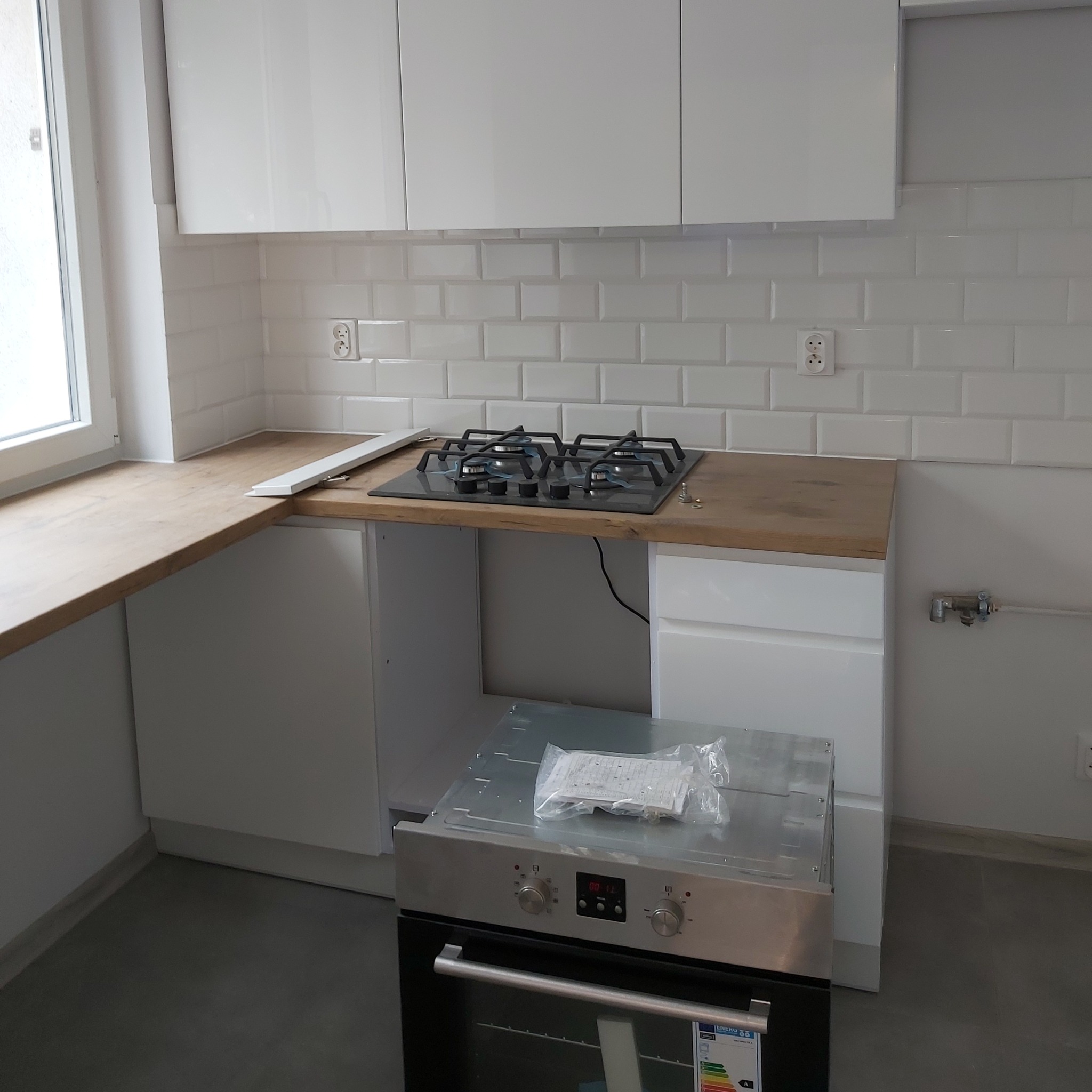 Montaż nowej płyty gazowej i piekarnika w kuchni z białymi szafkami i drewnianym blatem, widoczne białe płytki na ścianie.