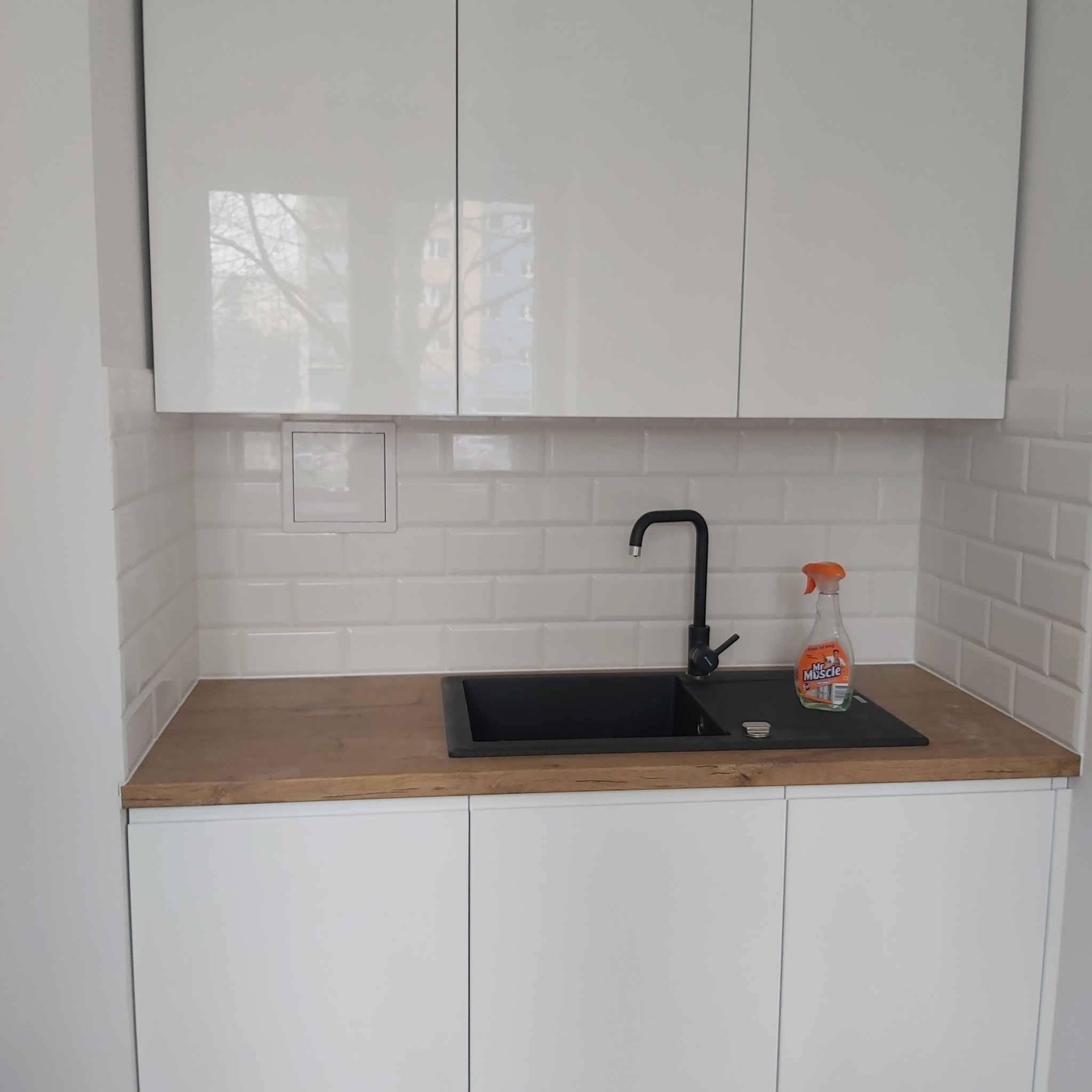 Biała kuchnia z czarnym zlewem i baterią, drewniany blat, białe kafelki typu metro, butelka środka czyszczącego Mr. Muscle stoi obok zlewu.