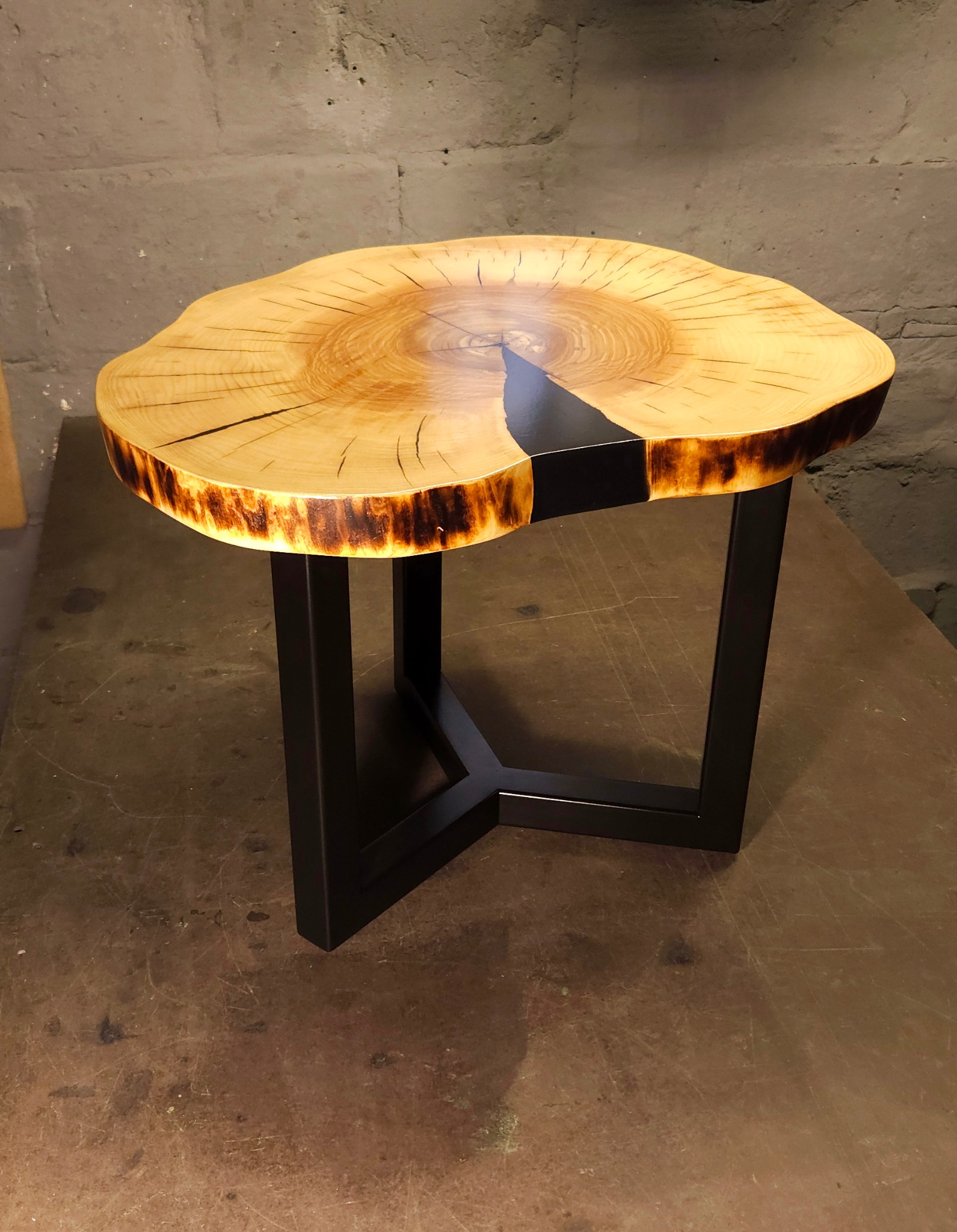 Stolik kawowy z blatem z plastra drewna wypełnionego czarną żywicą epoksydową, osadzony na metalowej, czarnej podstawie w kształcie litery X.