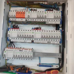 Rozdzielnica elektryczna w trakcie montażu, widoczne zabezpieczenia różnicowoprądowe, ograniczniki przepięć, przewody, listwy zaciskowe oraz narzędzia: śrubokręty i ołówki.