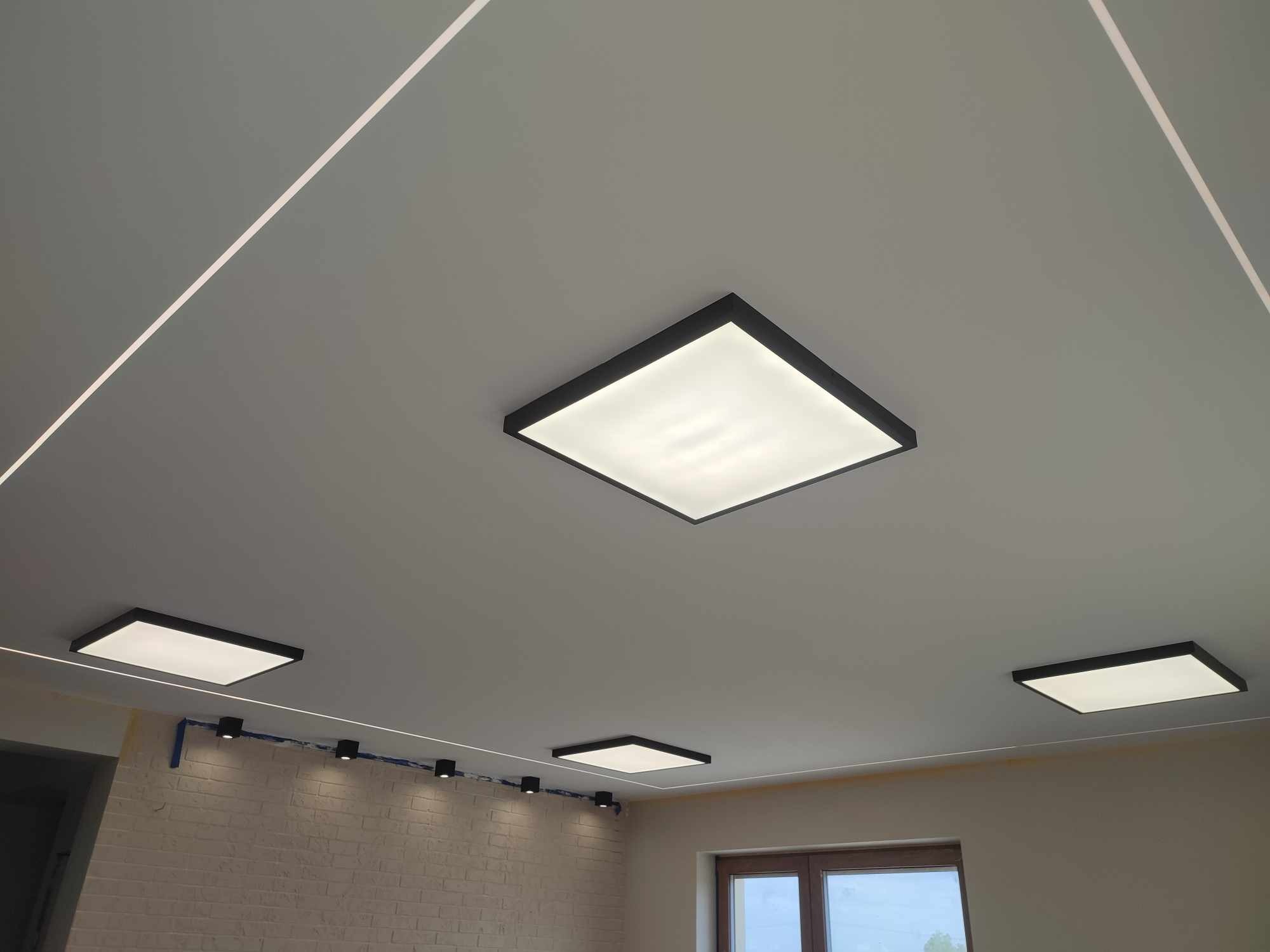 Zamontowane kwadratowe panele LED w czarnych ramkach na suficie, z widocznymi liniami LED i punktowym oświetleniem na ścianie z cegły.