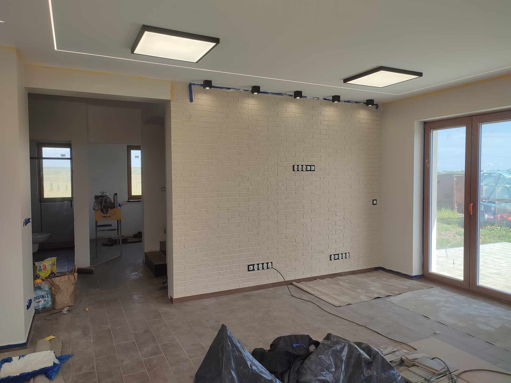 Wnętrze w trakcie wykańczania: ściana z białej cegły, zamontowane czarne, punktowe lampy sufitowe i kwadratowe panele LED, widoczne puszki elektryczne i kable, zabezpieczona podłoga, materiały...
