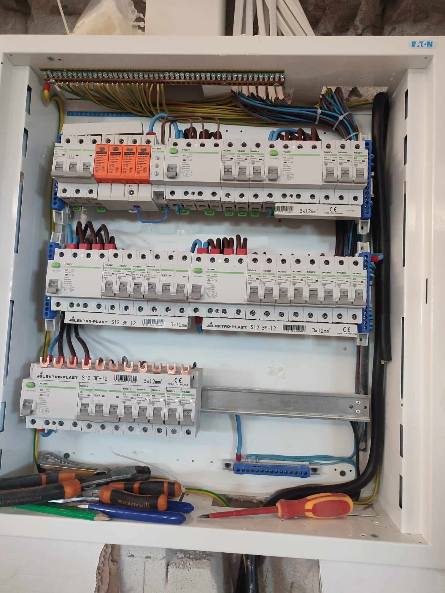 Rozdzielnica elektryczna w trakcie montażu, widoczne zabezpieczenia różnicowoprądowe, ograniczniki przepięć, przewody, listwy zaciskowe oraz narzędzia: śrubokręty i ołówki.