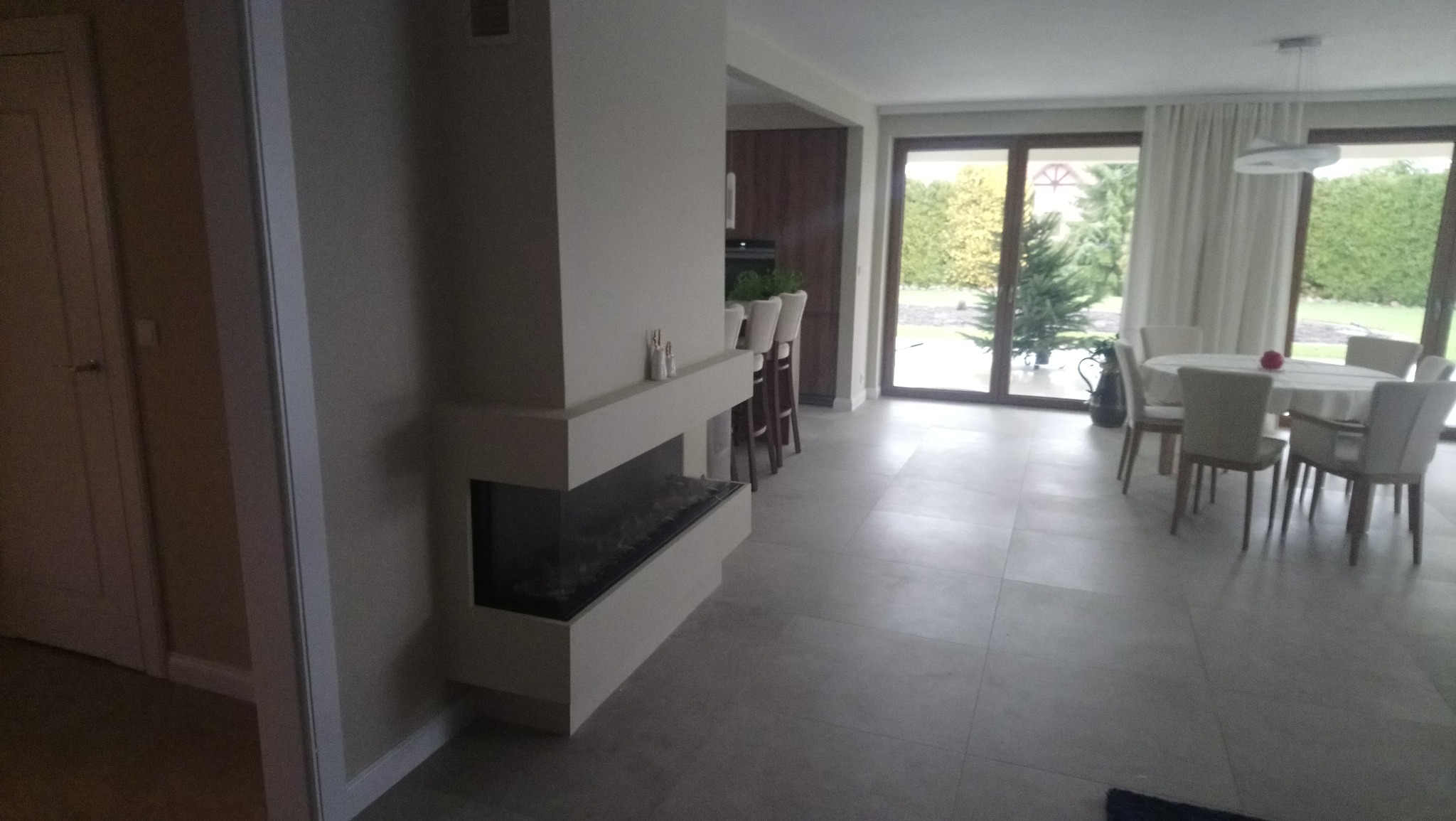 Nowoczesny salon z kominkiem narożnym i dużymi oknami. Jasne płytki podłogowe, stół z krzesłami w tle. Minimalistyczny design, neutralna kolorystyka.