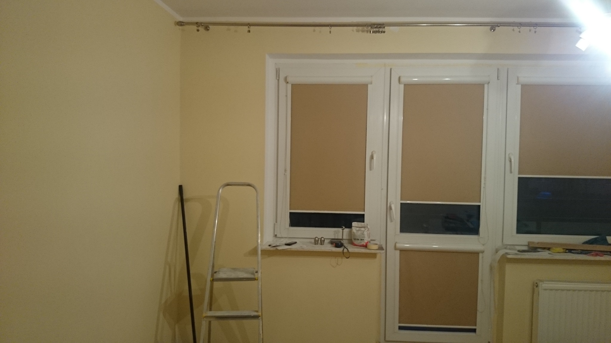 Pokój przed malowaniem: ściany w odcieniach beżu, okno z roletami, drabina oparta o ścianę, widoczne narzędzia malarskie na parapecie. Przygotowania do remontu.