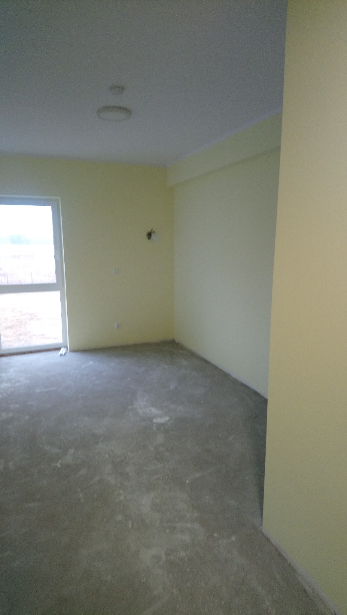 Pomalowane na jasnożółto wnętrze pokoju z oknem i surową podłogą. Widoczne ślady po pracach wykończeniowych, lampa na ścianie i sufitowe oświetlenie.