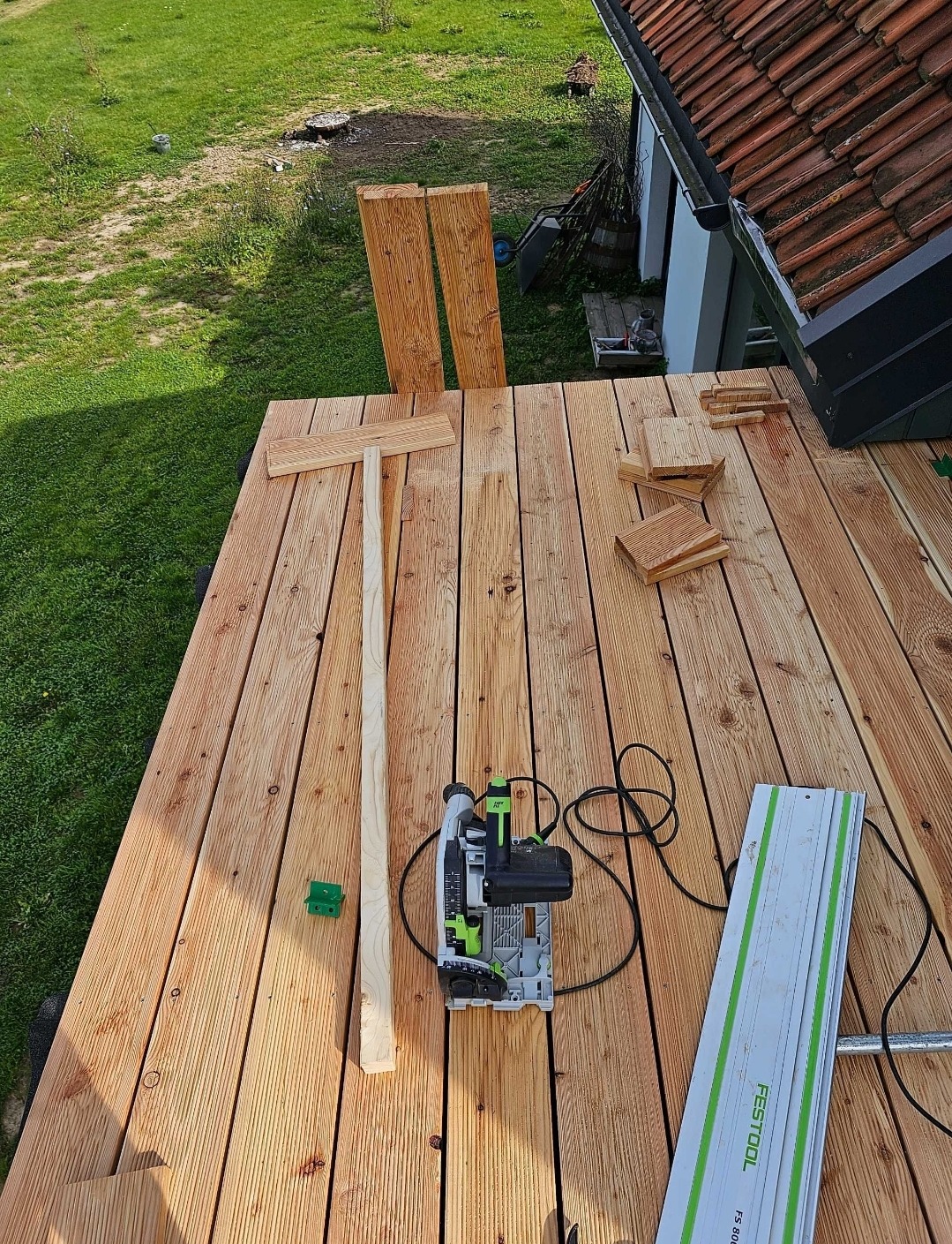 Montaż tarasu drewnianego: deski, piła elektryczna Festool, prowadnica i elementy konstrukcyjne na częściowo ułożonym tarasie, widok z góry na tle trawnika i dachu domu.
