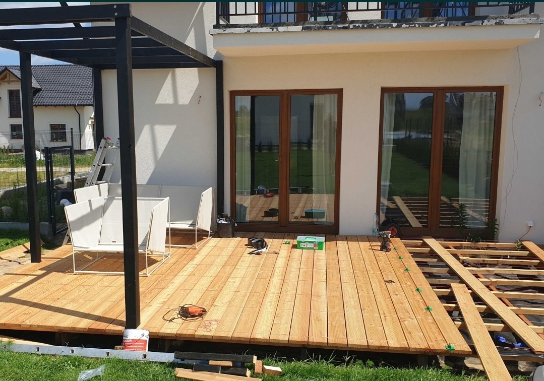 Budowa tarasu przy białym domu z dużymi oknami, widoczna konstrukcja nośna, deski tarasowe, narzędzia i elementy montażowe, czarna pergola z białymi meblami ogrodowymi w tle.