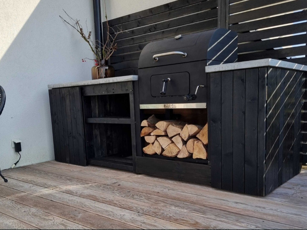 Czarna, drewniana zabudowa grilla na tarasie z ułożonym drewnem opałowym, z blatem z szarego kamienia i dekoracyjnym wazonem z gałązkami.