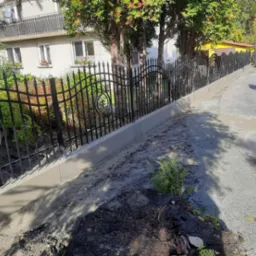 Czarny metalowy płot z ozdobnymi elementami na betonowej podmurówce, widok z boku na tle budynku i zieleni, fragment pnia drzewa w pierwszym planie.